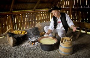 Mężczyzna w tradycyjnym stroju ludowym, z kapeluszem, stoi obok garnka z płynnym serem, przygotowywanego na ogniu. W jednej ręce trzyma kawałek sera, a drugą gestykuluje. W tle widać drewniane wnętrze chaty, a obok znajduje się drewniana deska z jedzeniem oraz kubełek z narzędziami. Na ziemi leżą kamienie.